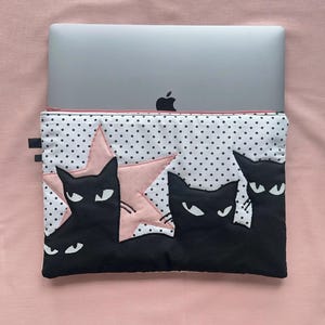以下が含まれることがあります： 黒猫のデザインのラップトップスリーブ。スリーブは、黒猫とピンクの星が付いた白い水玉模様です。ラップトップはシルバーで、スリーブの中に収まります。背景は無地のピンク色です。