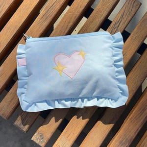 Peut inclure: Une pochette rectangulaire bleu clair avec un cœur rose et des étoiles dorées brodées. La pochette a un bord à volants et une fermeture éclair. L'accessoire est posé sur un banc en bois.
