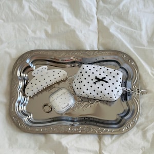 Könnte beinhalten: Ein silbernes Tablett enthält eine weiß-schwarze Haarklammer mit Polka-Dots, einen kleinen Beutel mit schwarzer Schleife und eine transparente Hülle mit dem Text "Round dot" für kabellose Ohrhörer. Das Tablett liegt auf einer weißen, strukturierten Oberfläche.