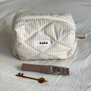May include: A beige and white striped quilted cosmetic bag with the name "Lulu" on a label. A tube of "Baby Got Bronze" bronzer and a gold-colored key are next to the bag. The bag has a zipper closure and a small handle.