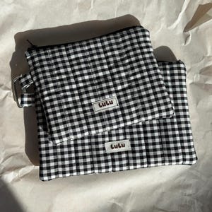 May include: Two black and white gingham pattern pouches. The larger pouch has a zipper closure and a small handle with a metal ring. Both pouches have a small rectangular tag that reads "Lulu".