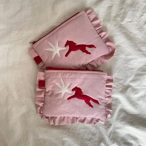 Puede incluir: Dos estuches rosas con cremallera, ribete rojo y bordes con volantes. Cada estuche presenta una estrella blanca y una aplicación de caballo rojo. Los estuches están hechos de tela suave.