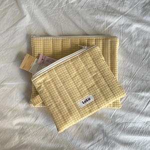 Pode incluir: Duas bolsas de cosméticos acolchoadas em xadrez amarelo e branco com zíperes brancos. A bolsa menor tem uma etiqueta que diz "Lulu". As bolsas estão em um lençol branco, e uma está parcialmente aberta, revelando pequenos tubos.