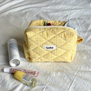 Op de afbeelding: Een gele geruite gewatteerde cosmetische tas met een witte rits en de naam "Lulu" op een wit label. De tas is open en onthult een paar items binnenin. Verschillende schoonheidsproducten zijn rond de tas gerangschikt.