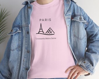 Puede incluir: Una camiseta rosa claro con un gráfico negro de la Torre Eiffel y la Pirámide del Louvre, con la palabra "PARIS" arriba y "Croissants.Métro.Seine" abajo. La camiseta se usa con una chaqueta vaquera azul.
