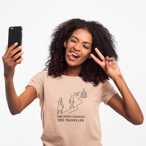 Puede incluir: Camiseta de color beige claro con el texto "THE PATH CHANGES THE TRAVELLER" y un gráfico de un camino, personas, una mariposa y una pagoda. La persona de la imagen sostiene un smartphone negro y hace el signo de la paz.