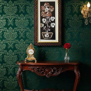 K&ouml;nnte beinhalten: Ein gerahmtes Kunstwerk mit einem floralen Arrangement aus Muscheln, &uuml;ber einem Holztisch. Eine goldene Uhr und eine Vase mit einer roten Rose stehen auf dem Tisch. Der Hintergrund ist eine dunkelgr&uuml;ne Tapete mit Muster.