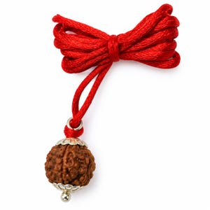 Könnte beinhalten: Ein brauner, kugelförmiger und texturierter Rudraksha-Perlenanhänger an einer leuchtend roten Kordel. Die Perle ist oben und unten mit silberfarbenen Metallverzierungen versehen, und die rote Kordel ist zu einer dekorativen Schleife gebunden.