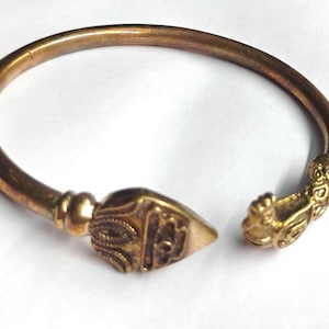 Pode incluir: Uma pulseira de punho aberta dourada. A pulseira apresenta um fecho em forma de ponta de flecha com detalhes gravados intrincados. A outra extremidade da pulseira tem um fecho decorativo com um design detalhado. A pulseira é feita de metal com um acabamento quente e antigo.