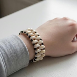 Könnte beinhalten: Ein cremefarbenes Muschelarmband mit schwarzen Perlen. Das Armband wird am Handgelenk getragen und zeigt die natürlichen Farbtöne der Muscheln und den Kontrast zu den schwarzen Perlen. Es ist ein Schmuckstück.
