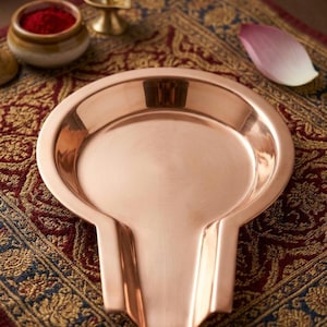 Pode incluir: Uma base yoni Shiva Lingam de cobre polido com uma bacia circular e um bico alongado proeminente que se estende pela frente. A superfície metálica tem um acabamento quente e reflexivo, apresentando um design liso e limpo para uso ritual.