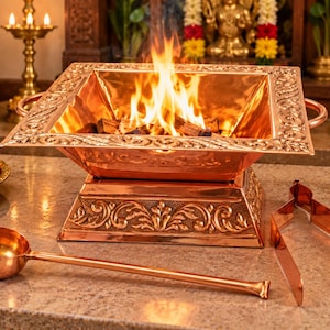 Puede incluir: Un brasero cuadrado de cobre con un borde y una base ornamentados con motivos florales en relieve, que contiene una llama naranja brillante. Un cucharón de cobre de mango largo y unas pinzas de cobre se sitúan frente al brasero sobre piedra.