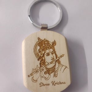 Peut inclure: Un porte-clés en bois clair avec une illustration gravée au laser représentant une figure portant une couronne ornée et jouant de la flûte. Le texte "Shree Krishna" est gravé sous la figure. La pièce en bois est fixée à un anneau en métal argenté.