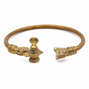 May include: A gold-toned metal open cuff bracelet featuring a smooth, rounded band. One end terminates in a detailed sculpted head, while the other end features a sculpted head adjacent to a small, upright cobra figure atop a pedestal base.