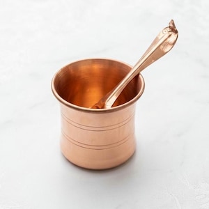 May include: A copper-colored cup with a matching spoon. The cup has three horizontal lines etched around its body. The spoon rests inside the cup, with its handle extending out. The items are set against a white background.