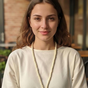 May include: A woman wearing a cream-colored top and a long beaded necklace with a red tassel. The necklace is made of small, round, light-colored beads. The woman has long, wavy brown hair and is smiling.