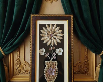 Könnte beinhalten: Ein gerahmtes Kunstwerk mit einem Blumenarrangement aus Muscheln. Das Arrangement umfasst eine große Blume, kleinere Blüten und eine Vase, die alle aus verschiedenen Muscheln hergestellt wurden. Der Rahmen ist dunkelbraun mit einem goldenen Innenrand. Der Hintergrund ist schwarz.