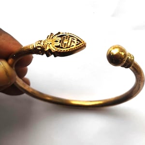 Puede incluir: Una pulsera de brazalete de color dorado con un diseño abierto. Un extremo presenta un elemento decorativo y ornamentado, mientras que el otro extremo tiene un acento redondo en forma de bola. La pulsera se sostiene sobre un fondo blanco.