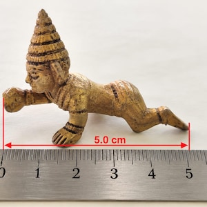 Puede incluir: Una pequeña figura de tono dorado que representa a una persona gateando con un sombrero puntiagudo y texturizado. La figura está colocada sobre una regla metálica, mostrando una longitud total de 5,0 cm según las líneas de medición rojas.