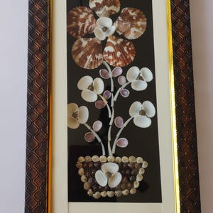 May include: Framed artwork featuring a floral arrangement crafted from seashells. The flowers are white and brown, set against a black background. The frame is brown with a gold inner border, creating a decorative piece.