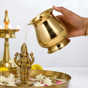 May include: A polished brass vessel pouring water over a small brass deity figurine. The figurine stands on a circular brass tray decorated with white flowers and pink petals. A brass oil lamp stands in the background, completing the scene.