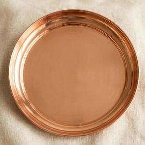 May include: A circular copper tray with a polished, reflective surface and a smooth, raised rim. The metal has a warm, reddish-brown metallic tone with subtle horizontal light reflections across the center of the flat, round base.