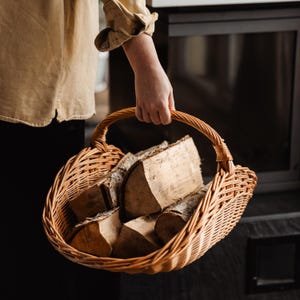 May include: A woven wicker basket filled with firewood. The basket has a curved handle and is held by a person wearing a tan shirt. The firewood pieces are various sizes and shapes, with visible bark and wood grain.