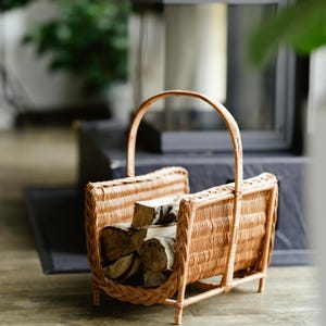 Puede incluir: Una cesta de mimbre marrón claro, llena de leña cortada. La cesta tiene un asa curva y una forma rectangular con un diseño tejido. La leña está formada por varios trozos de madera. La cesta está hecha de mimbre tejido.