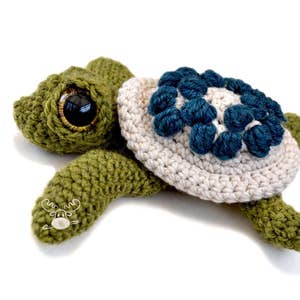 K&ouml;nnte beinhalten: Handgefertigte H&auml;kelschildkr&ouml;te in Gr&uuml;n, Creme und Blau. Die Schildkr&ouml;te hat gro&szlig;e, ausdrucksstarke Augen und einen Panzer, der mit blauen Blumenmotiven verziert ist. Eine charmante Dekoration.