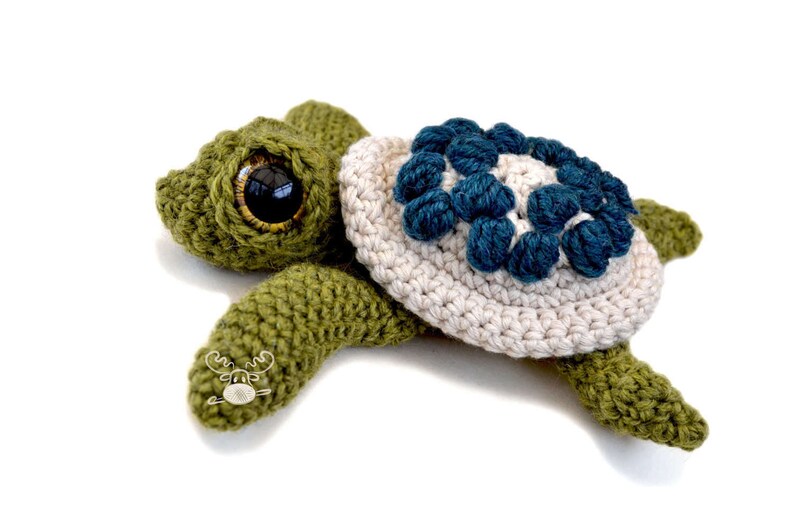 K&ouml;nnte beinhalten: Handgefertigte H&auml;kelschildkr&ouml;te in Gr&uuml;n, Creme und Blau. Die Schildkr&ouml;te hat gro&szlig;e, ausdrucksstarke Augen und einen Panzer, der mit blauen Blumenmotiven verziert ist. Eine charmante Dekoration.