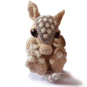 Puede incluir: Un juguete de peluche de armadillo tejido a crochet con pelaje marrón y blanco. El armadillo tiene un hocico pequeño y puntiagudo y ojos grandes y marrones.