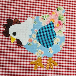 May include: A colourful fabric chicken applique. The chicken features a white head, brown comb, and yellow beak. The body is light blue with floral and plaid patterns, set against a red and white gingham background.