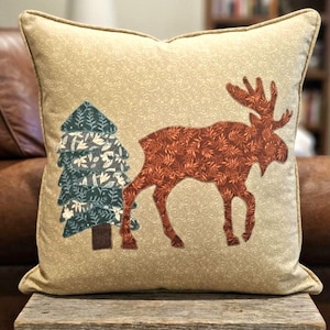 May include: Square decorative pillow with a beige background and a repeating swirl pattern. The pillow features an applique of a brown moose and a blue and green tree. The pillow has a beige border.