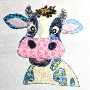 Puede incluir: Aplicación de vaca de tela colorida con un diseño de patchwork. La vaca presenta una nariz floral rosa, orejas estampadas azules y blancas y una corona. El fondo es blanco.