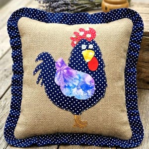 May include: Square burlap pillow with a blue polka dot border and a ruffled edge. The pillow features a whimsical rooster applique with a red comb, yellow beak, and a purple and blue body. The rooster is set against a blue polka dot background.