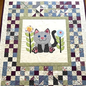 Puede incluir: Edredón hecho a mano con un gato gris con lunares y flores. El borde es de patchwork en tonos azules, morados y crema. El edredón está cosido a mano, creando un diseño caprichoso y acogedor.