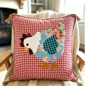 Pode incluir: Almofada quadrada decorativa com padrão xadrez vermelho e branco. A almofada apresenta uma aplicação de tecido de uma galinha com cabeça branca, corpo azul e detalhes florais. A almofada tem uma borda de corda marrom e borlas.
