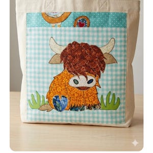 May include: A beige tote bag with a colorful appliqué of a Highland cow. The cow has a shaggy brown coat, large horns, and is set against a blue and white gingham pattern. The bag has sturdy beige handles.