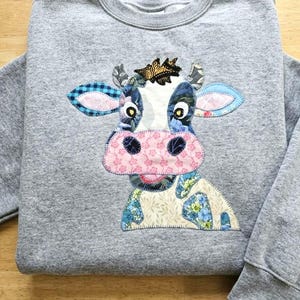 Puede incluir: Una sudadera gris con un diseño de vaca de patchwork. La vaca tiene una nariz rosa, orejas estampadas en azul y blanco y una corona. El diseño está hecho de varios patrones de tela, incluyendo florales y cuadros.