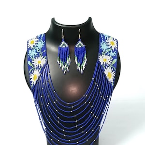 May include: A beaded necklace and earring set displayed on a black mannequin. The necklace is primarily blue with white and yellow floral accents and cascading beaded strands. The matching earrings are blue with white and green fringe.