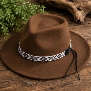 May include: A brown fedora hat featuring a wide brim and a pinched crown. The hat is accented with a white beaded hat band displaying a geometric diamond pattern in black and brown tones, finished with a black leather side tab and thin ties.