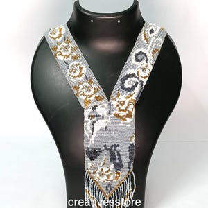 May include: A beaded necklace with a gray background, featuring white, gold, and black floral and abstract patterns. The necklace has a V-shaped design with a central pendant and a fringe of beads at the bottom. The text "creativesstore" is visible.