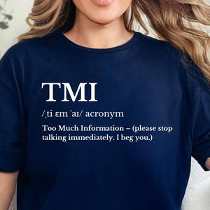 May include: Navy blue t-shirt with the acronym "TMI" in white, along with its pronunciation and definition: "Too Much Information - (please stop talking immediately. I beg you.)". The shirt is worn with light blue ripped jeans.