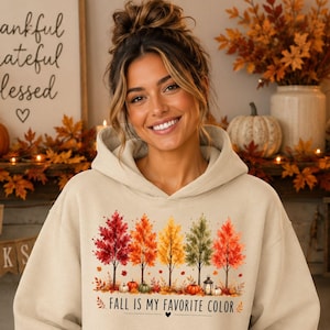 May include: A beige hooded sweatshirt featuring a graphic print of five colorful autumn trees in shades of red, orange, and yellow. Below the trees are small pumpkins and a lantern, with the text "FALL IS MY FAVORITE COLOR" printed in black.