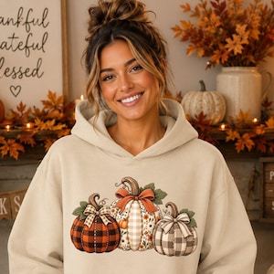 May include: A beige hooded sweatshirt featuring a graphic print of three pumpkins. The left pumpkin has a red and black buffalo plaid pattern, the center pumpkin has a floral and beige gingham design with a bow, and the right pumpkin has a gray gingham pattern.