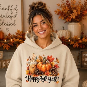 May include: A beige hooded sweatshirt featuring a graphic print of an iced coffee, a pumpkin, pinecones, and autumn leaves. The text "Happy Fall Y'all" is printed in a black script font below the colorful seasonal illustration.