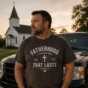 May include: A dark brown t-shirt with the text "FATHERHOOD IS KINGDOM WORK" in white, along with a cross and the words "LEAD LOVE THAT LASTS" and "PROVERBS 22:6". A man is wearing the shirt, standing in front of a church and a black truck.