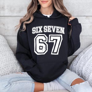 May include: Black hooded sweatshirt with the text "SIX SEVEN" above the number "67" in white, outlined in black. The sweatshirt is worn by a person sitting down.