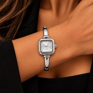 May include: A silver-toned quartz wristwatch featuring a square face with a minimalist silver dial, thin hour and minute hands, and a polished metal bangle-style strap that connects directly to the top and bottom of the watch case.