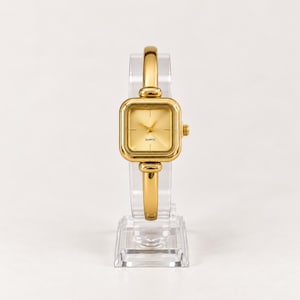 May include: A gold-toned analogue wristwatch featuring a square case with rounded corners and a matching bangle-style band. The dial is gold-coloured with simple hour markers and the word QUARTZ printed in black text below the centre hands.
